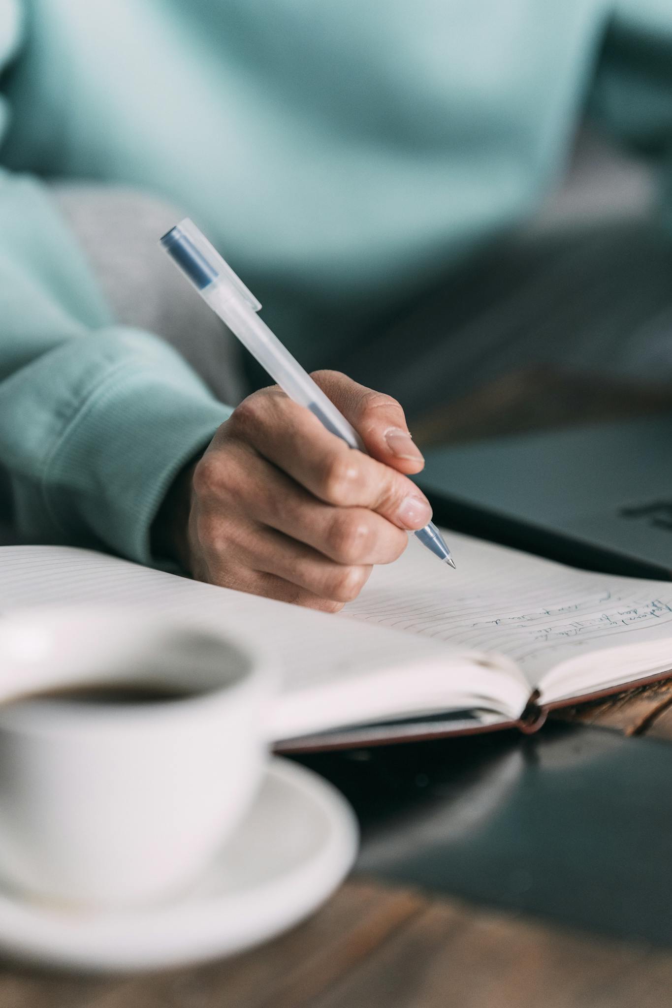 Close-up of a person writing in a notebook with coffee and laptop nearby, indoors.